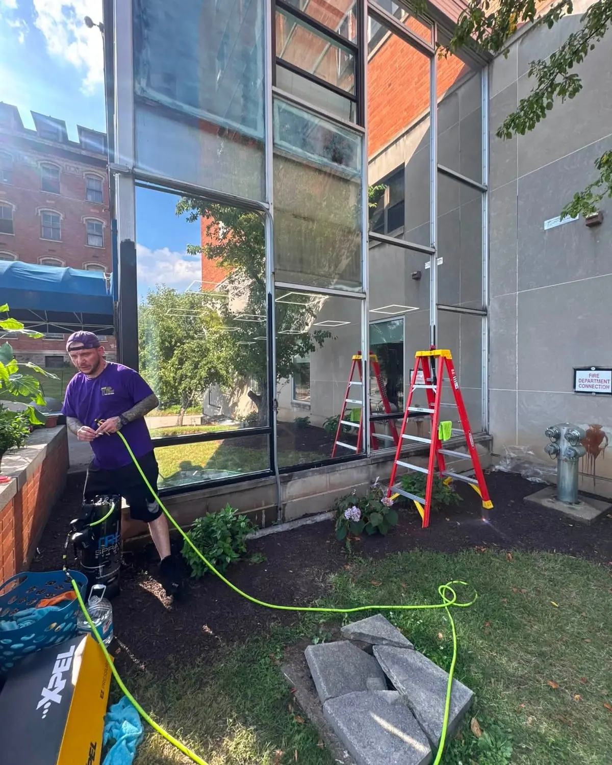 A Mr. Tint installer applying window film to a commercial window, with a ladder and tools beside him