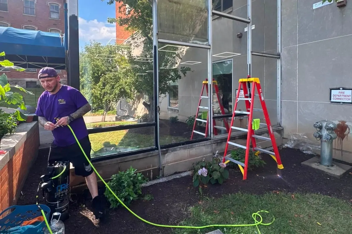 A Mr. Tint installer applying window film to a commercial window, with a ladder and tools beside him